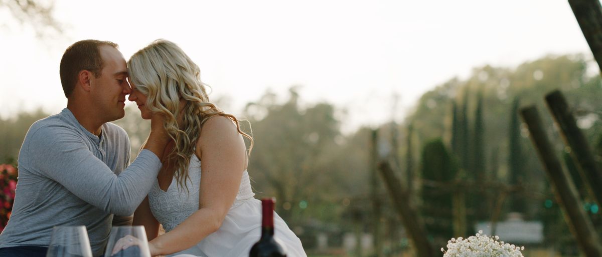 Taylor and Alec resting their foreheads together at a vineyard table